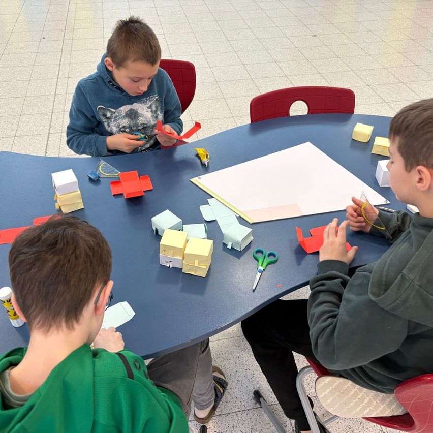 Drei Jungen sitzen um einen Tisch herum und arbeiten an einem Projekt. Einer hält eine Schere, ein anderer Papier und der dritte konzentriert sich auf eine Papierkreation. Der Tisch ist mit Papieren, Scheren und kleinen gelben Objekten übersät.