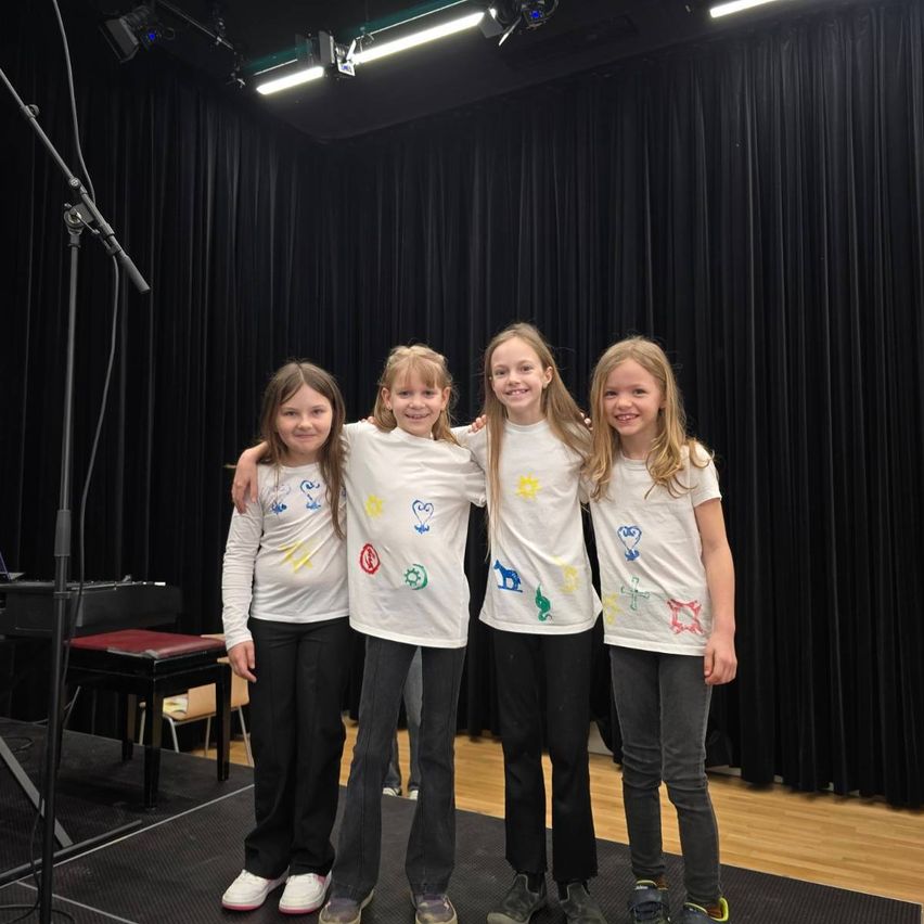 Vier Mädchen posieren für ein Foto in einem Studio mit schwarzen Vorhängen, einem Klavier und einem Mikrofon. Sie tragen weiße T-Shirts mit bunten Zeichnungen.