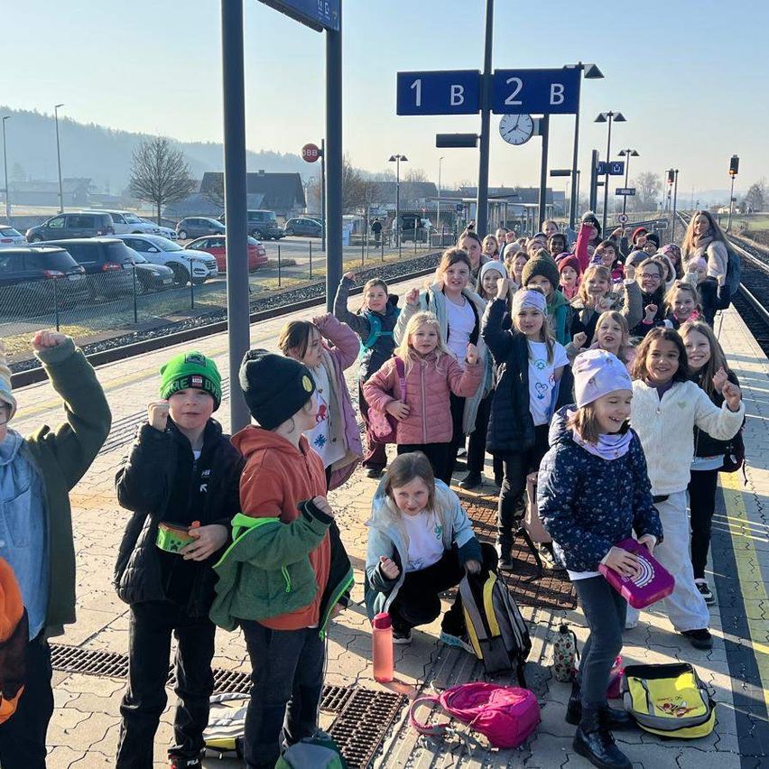 Eine Gruppe von Kindern mit ihren Lehrern steht auf einem Bahnsteig, hält ihre Taschen und posiert für ein Foto. Der Bahnsteig hat eine Uhr, und ein Stoppschild ist auf dem Pfosten. Der Parkplatz ist hinter ihnen, und die Bahnlinie ist rechts.