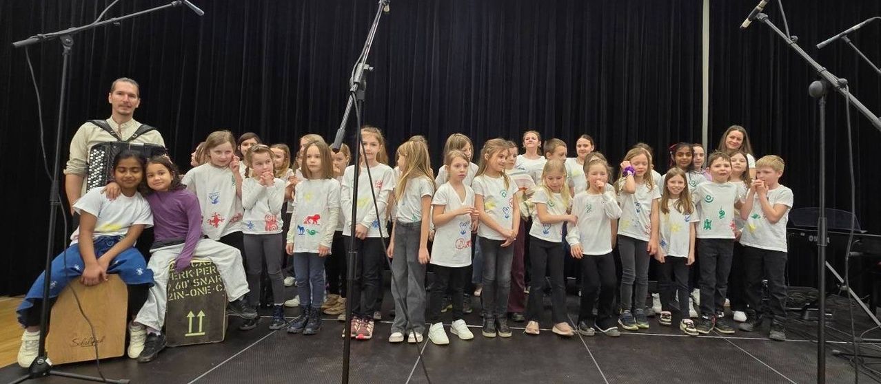Eine Gruppe junger Mädchen in weißen Shirts steht auf einer Bühne, wahrscheinlich für eine Aufführung. Sie halten Händchen und scheinen für ein Foto zu posieren.