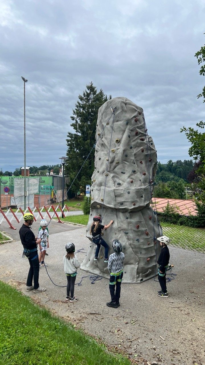 Eine Gruppe von Kindern und Erwachsenen klettert an einer Kletterwand. Die Kinder tragen Helme und Gurte. Die Erwachsenen beaufsichtigen. Bäume und ein Gebäude sind im Hintergrund.