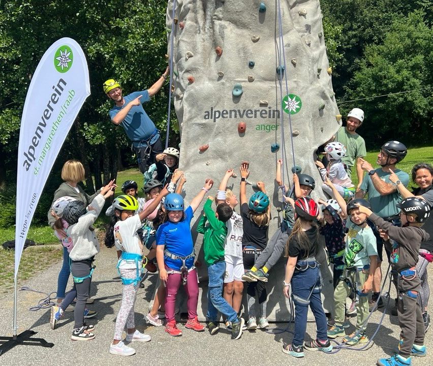 Eine Gruppe von Kindern und Erwachsenen klettert gemeinsam, trägt Helme und Gurte. Die Kletterwand hat den Text 'alpenverein angel' darauf.