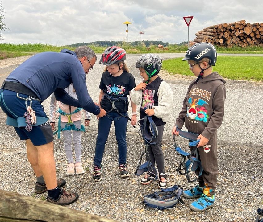 Ein Ausbilder zeigt einer Gruppe von Kindern draußen, wie man Kletterausrüstung anlegt. Sie tragen alle Helme.