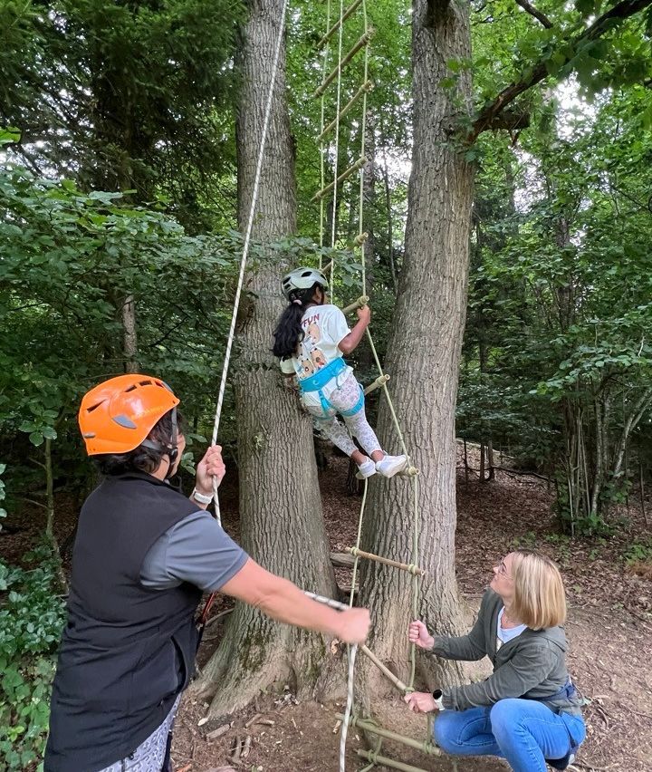 Ein Mädchen klettert eine Seilleiter an einem Baum hoch, während ein Mann und eine Frau beobachten. Der Mann trägt einen Helm und hält das Seil, während die Frau zusieht und ein Seil hält.