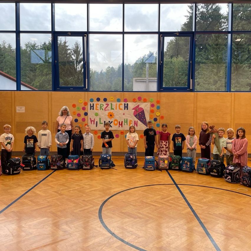 Eine Gruppe von Kindern und einer erwachsenen Frau stehen in einer Turnhalle mit Rucksäcken, ein Banner mit der Aufschrift 'Herzlich Willkommen' im Hintergrund.