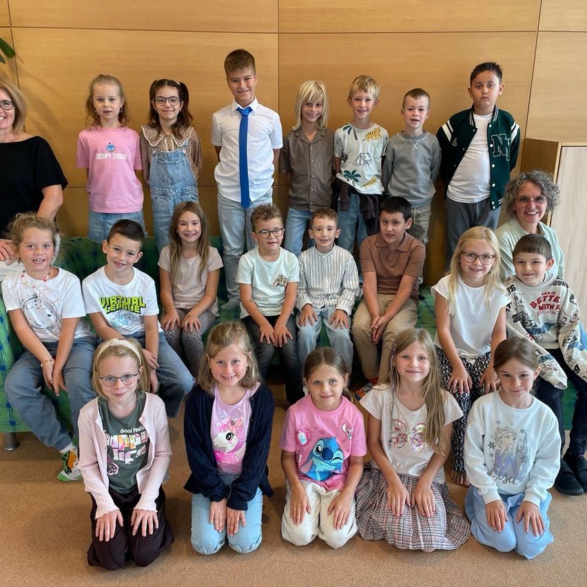 Eine Gruppe von Kindern und zwei Erwachsenen posiert für ein Foto in einem Raum mit einer Holzwand und einer grünen Couch.
