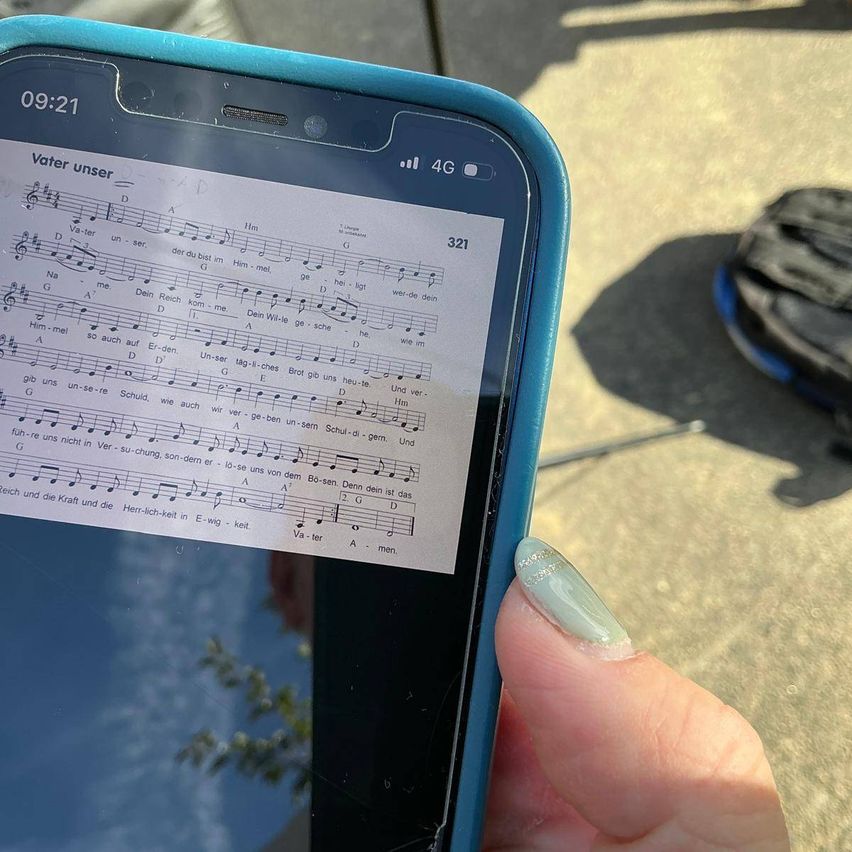 Eine Person hält ein Smartphone mit einem musikalischen Notenblatt, das 'Vater unser' beschriftet ist. Der Bildschirm zeigt Musiknoten und Texte, die Zeit wird als 09:21 angezeigt. Das Telefon ist in einem blauen Gehäuse.