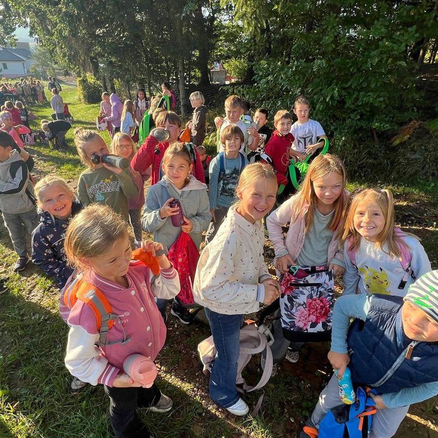 Eine Gruppe von Kindern, einige mit Wasserflaschen, steht auf einem Rasen mit Bäumen im Hintergrund. Sie lächeln und scheinen auf einem Schulausflug zu sein.