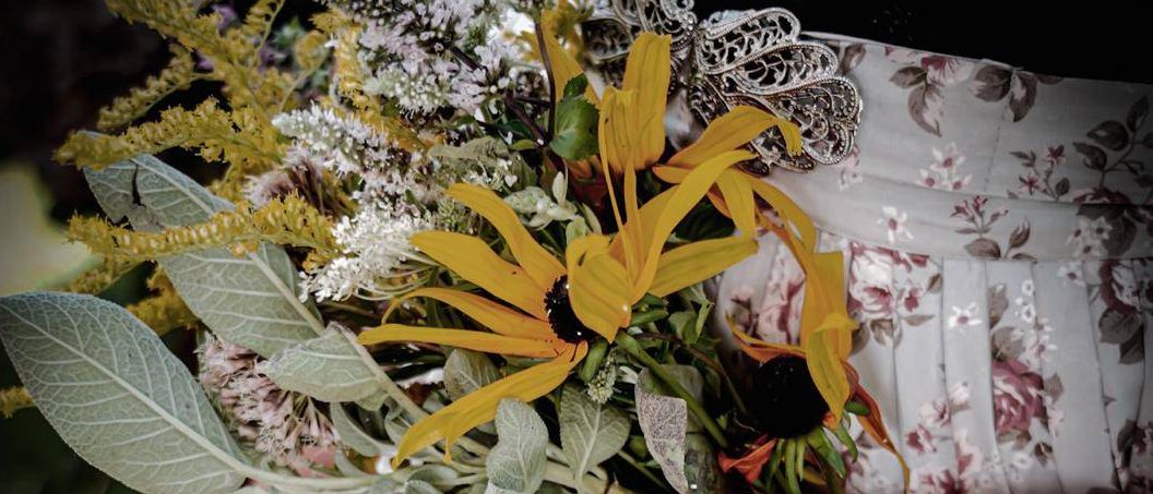 Ein Nahaufnahme der Hände einer Person, die einen Strauß gelber Sonnenblumen und grüner Blätter hält. Die Person trägt ein Blumenkleid.