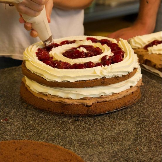 Eine Person dekoriert einen mehrschichtigen Kuchen mit Schlagsahne und Kirschen auf einer Marmorplatte.