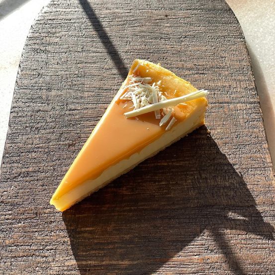 Ein Stück Karamell-Käsekuchen mit einem Karamell-Topping und weißen Schokoladenraspeln auf einem Holzbrett.