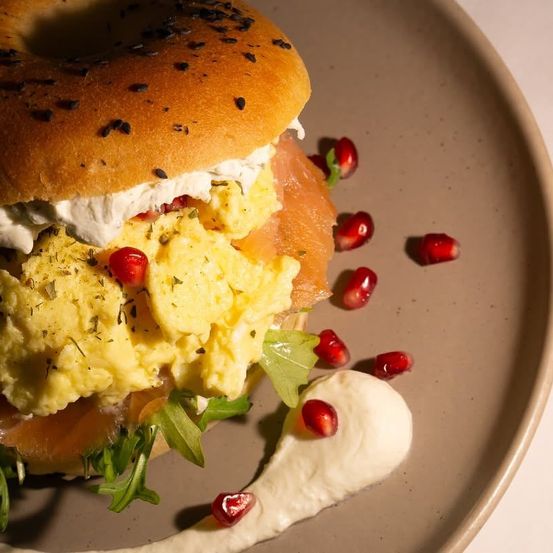Ein Bagel mit Rührei, Räucherlachs und Granatapfelsamen auf einem Teller mit einer cremigen Beilage.