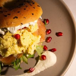 Ein Bagel mit Rührei, Räucherlachs und Granatapfelsamen auf einem Teller mit einer cremigen Beilage.