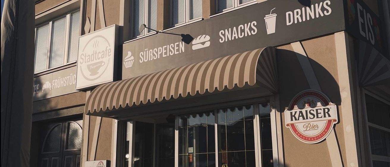 Ein Gebäude mit einem braunen Vordach zeigt Schilder für Stadtcafe, das Frühstück und Kaffee anbietet. Ein weiteres Schild bewirbt Snacks und Getränke. Es gibt Glasfenster und Türen, und ein Regenschirm steht links.