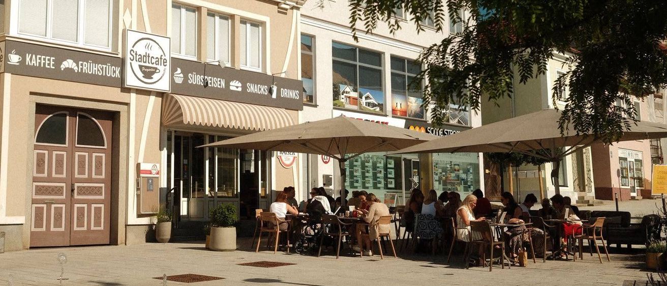 Ein Café im Freien mit großen Sonnenschirmen, Leuten, die an Tischen sitzen, einem Gebäude mit dem Schild 'Stadtcafe' und einem Baum im Vordergrund.