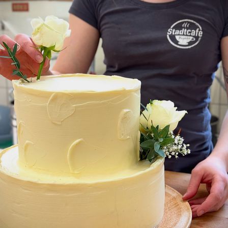 Eine Person verziert einen zweistöckigen Kuchen mit weißen Rosen und Blättern auf einem Holztisch. Der Kuchen hat eine glatte, cremefarbene gelbe Glasur.