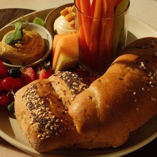 Ein Teller mit zwei Brotsorten, einem Glas Karottensticks, einer Schüssel Hummus und einer Schüssel mit verschiedenen Früchten.