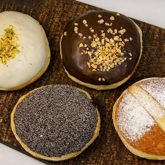Vier Donuts auf einem Holzbrett: weiß mit Pistazie, Schokolade mit Nüssen, schwarzer Sesam und Puderzucker.