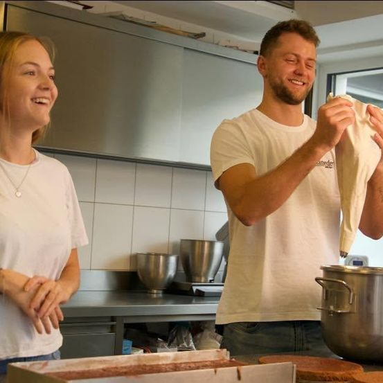 Ein Mann und eine Frau sind in einer Küche. Der Mann hält ein Tuch und lächelt, während die Frau ihn beobachtet. Auf dem Tresen befinden sich Schüsseln, ein Topf und eine Schachtel.