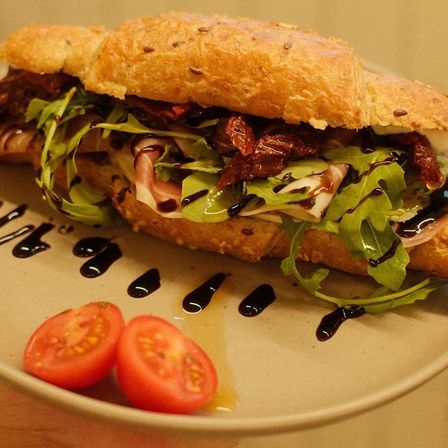Ein Sandwich mit geröstetem Brot, Rucola, getrockneten Tomaten und Balsamico-Drizzle auf einem weißen Teller mit zwei Kirschtomaten.