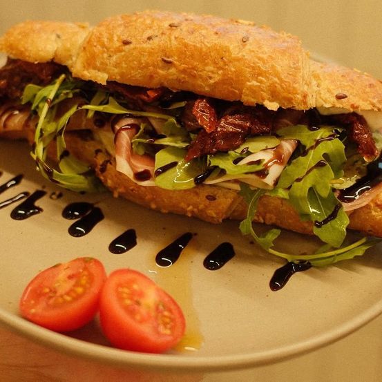 Ein Sandwich mit geröstetem Brot, Rucola, getrockneten Tomaten und Balsamico-Drizzle auf einem weißen Teller mit zwei Kirschtomaten.