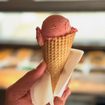 Eine Hand hält einen Erdbeereisbecher in einem Waffelkegel, mit einer Serviette darunter. Der Hintergrund ist ein verschwommener Eisladen.