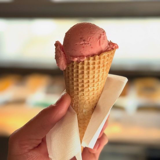 Eine Hand hält einen Erdbeereisbecher in einem Waffelkegel, mit einer Serviette darunter. Der Hintergrund ist ein verschwommener Eisladen.