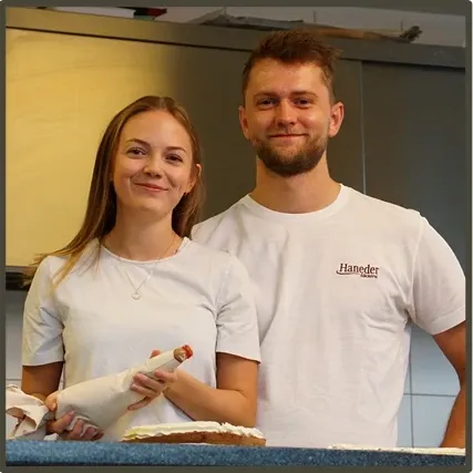 Ein Mann und eine Frau in weißen Haneder-T-Shirts stehen lächelnd und posieren gemeinsam in einer Küche für ein Foto. Die Frau hält eine Konditorblase, und ein Kuchen steht auf dem Tresen.
