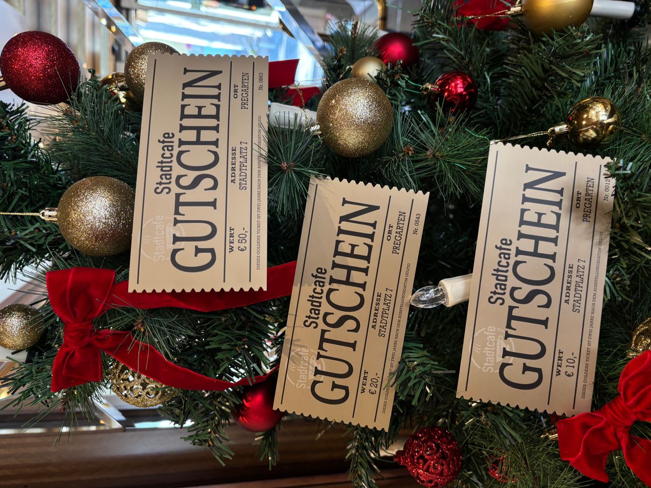 Ein Weihnachtsbaum, geschmückt mit Geschenkgutscheinen, die jeweils 'Gutscheine' mit den Werten von 60 € und 20 € tragen, ist mit goldenen und roten Kugeln dekoriert. Der Baum hat ein festliches Band und steht vor einem unscharfen Hintergrund.