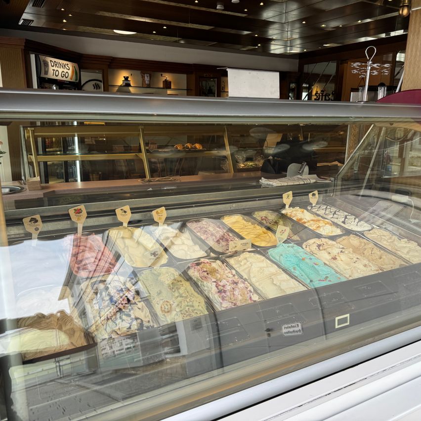 Eine Vitrine mit verschiedenen Eissorten. Im Hintergrund ist ein Schild mit der Aufschrift 'Drinks to go' zu sehen.