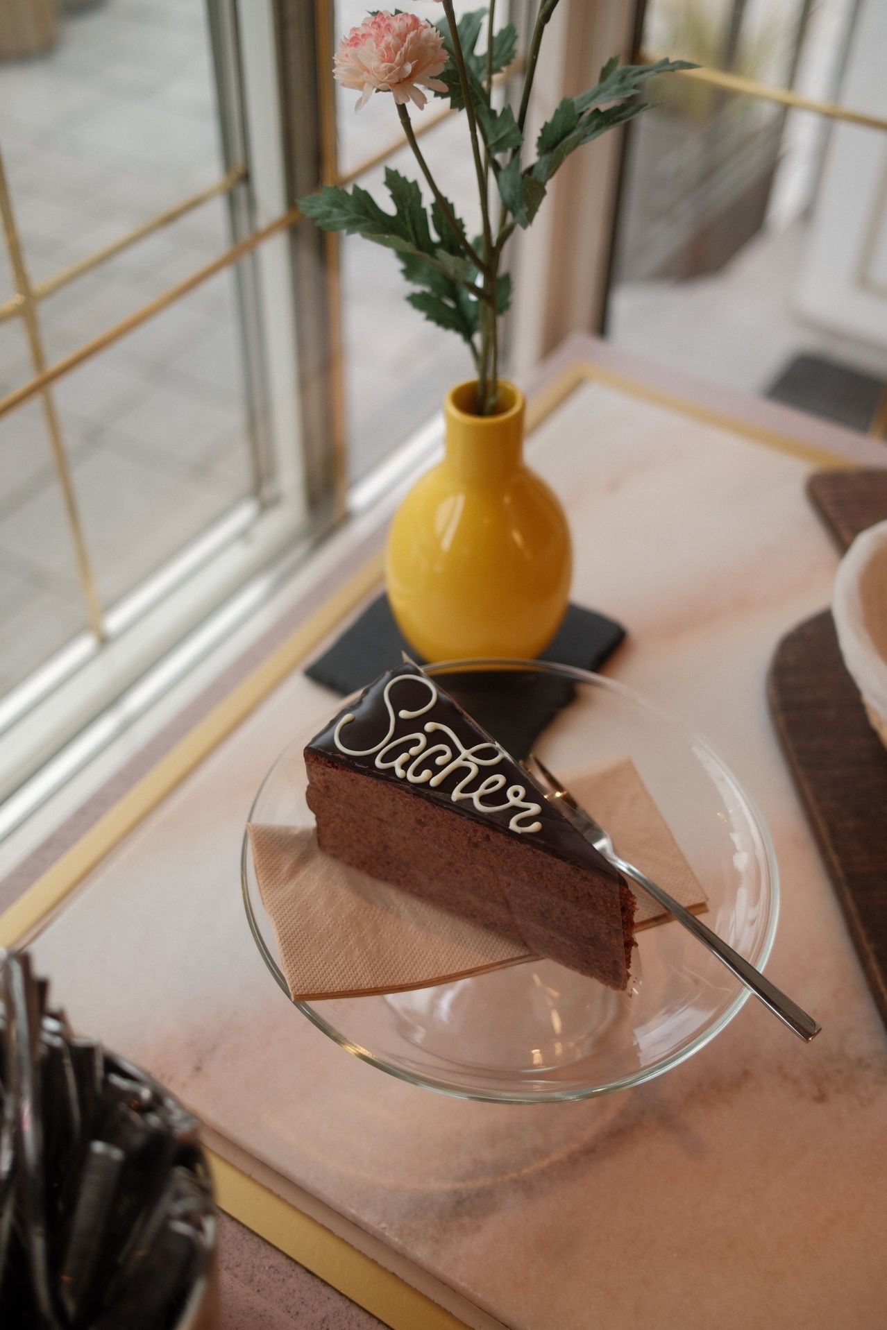 A slice of chocolate cake with 'Sacher' written in white icing sits on a glass plate with a fork. A yellow vase with a plant is behind it.