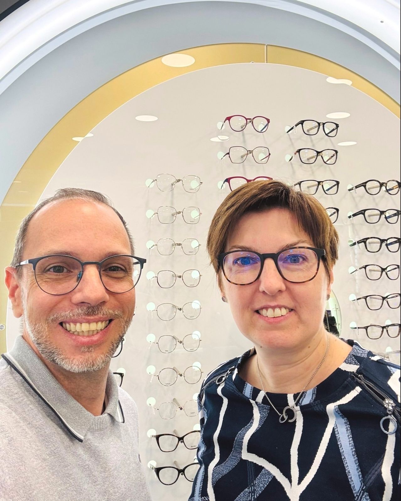 Ein Mann und eine Frau mit Brille lächeln und posieren vor einer Wand, auf der verschiedene Brillen ausgestellt sind.