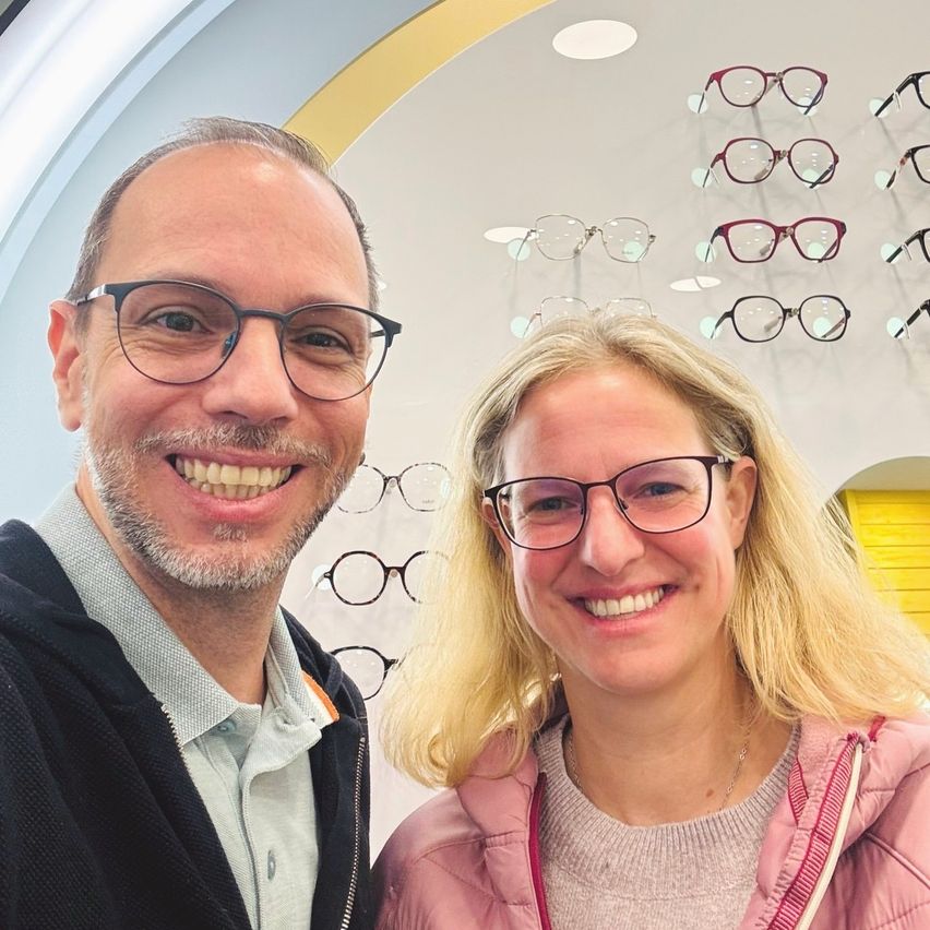 Ein Mann und eine Frau lächeln für ein Selfie vor einer Optikerwand, auf der verschiedene Brillenrahmen ausgestellt sind.