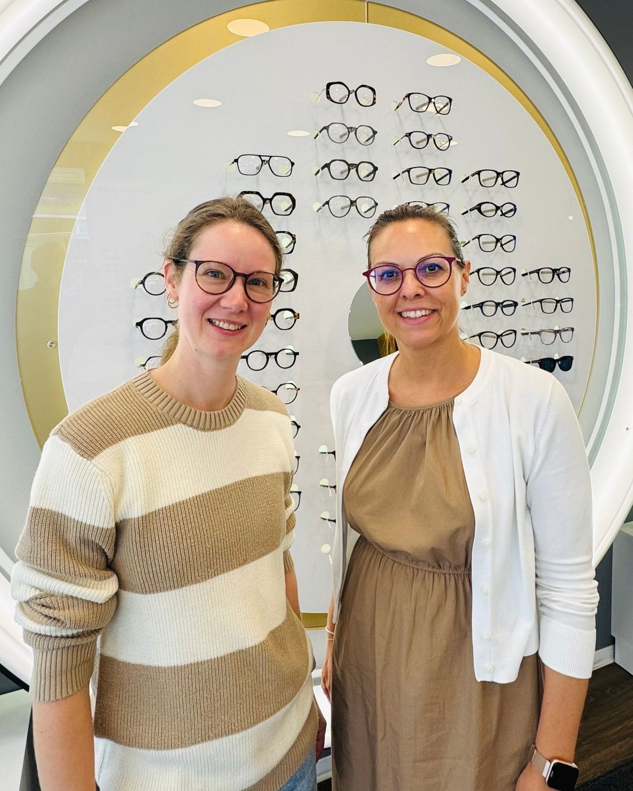 Zwei Frauen stehen lächelnd vor einer Wand mit mehreren Brillenpaaren.