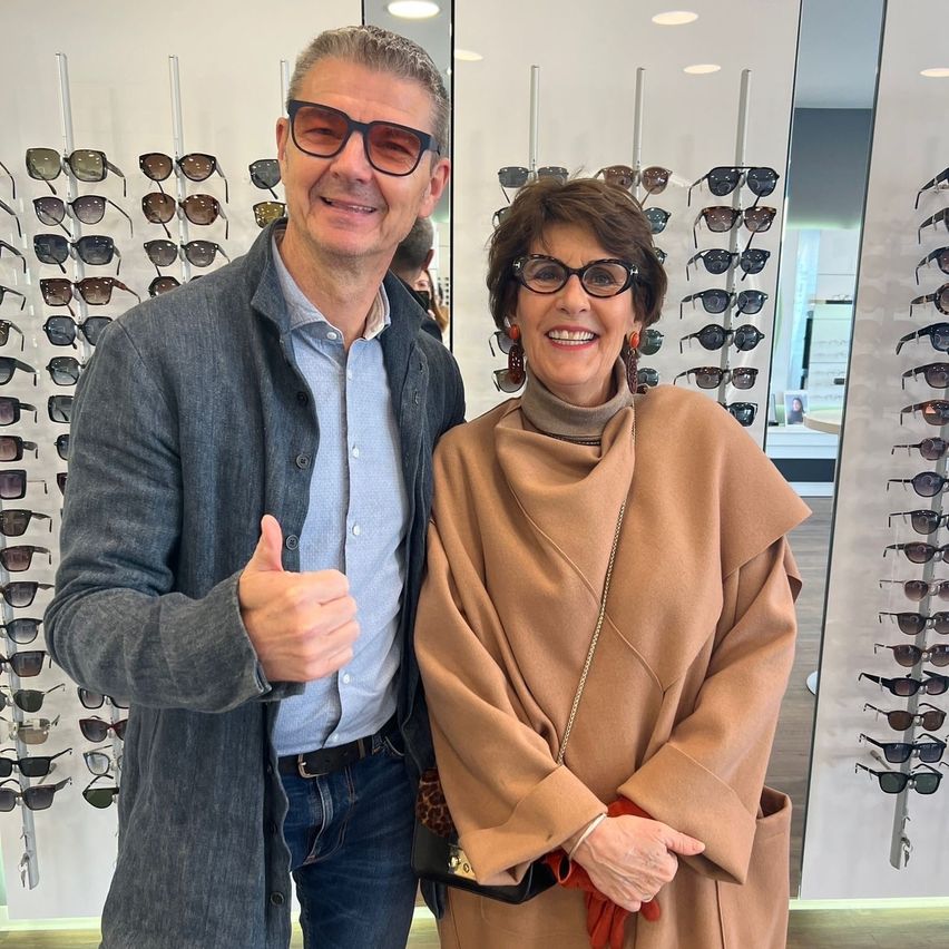 Ein Mann und eine Frau stehen lächelnd vor einer Wand voller Sonnenbrillen und posieren für ein Foto.
