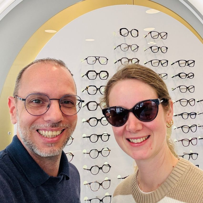 Ein Mann und eine Frau lächeln und posieren für ein Foto vor einer Wand mit vielen ausgestellten Brillen.