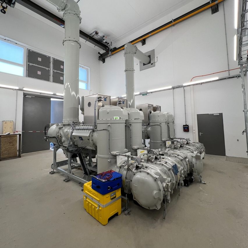 Ein Industriegebäude mit mehreren großen grauen Maschinen, einem gelben Lagerbox und einer grauen Tür. Der Raum ist gut beleuchtet mit weißen Wänden und Metallgeländern.