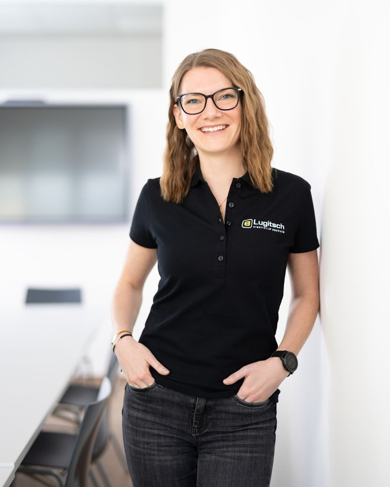 Eine Frau mit einer schwarzen Bluse und einem Logo auf der linken Brustseite lächelt, während sie vor einer weißen Wand steht.