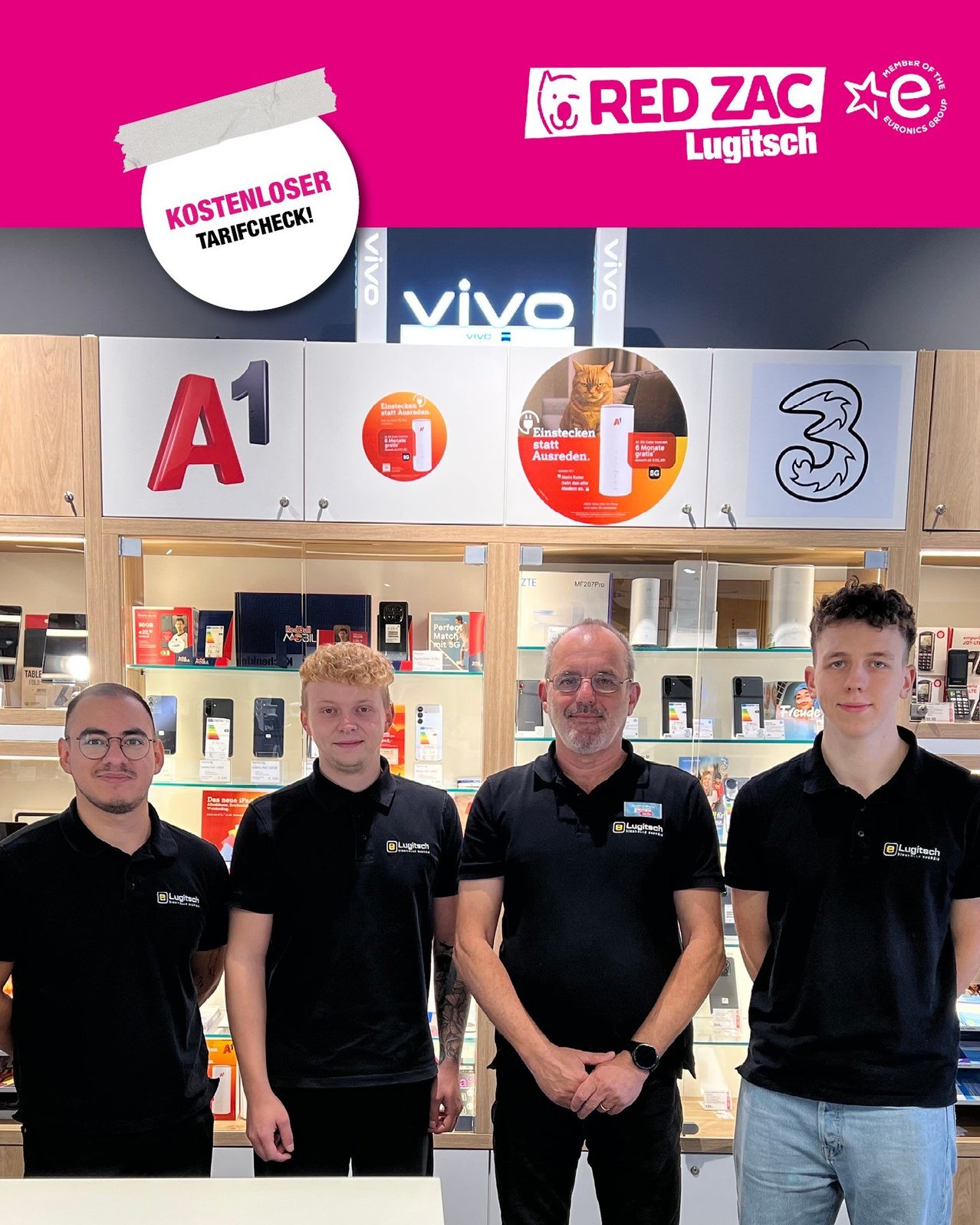 Vier Männer stehen vor einem Laden mit verschiedenen Elektronikgeräten. Sie tragen alle schwarze Poloshirts mit einem Logo. Hinter ihnen befinden sich Regale mit Elektronikartikeln. Auf den Regalen befinden sich drei Schilder mit Zahlen und Logos.