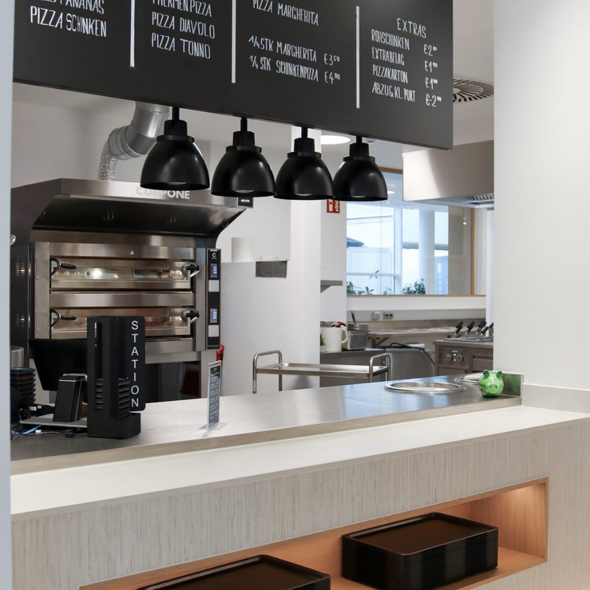 Eine moderne Küche mit einer Menütafel über einer Theke, die Pizza-Optionen und Extras anbietet, zusammen mit einem Edelstahlofen und einem Waschbecken.