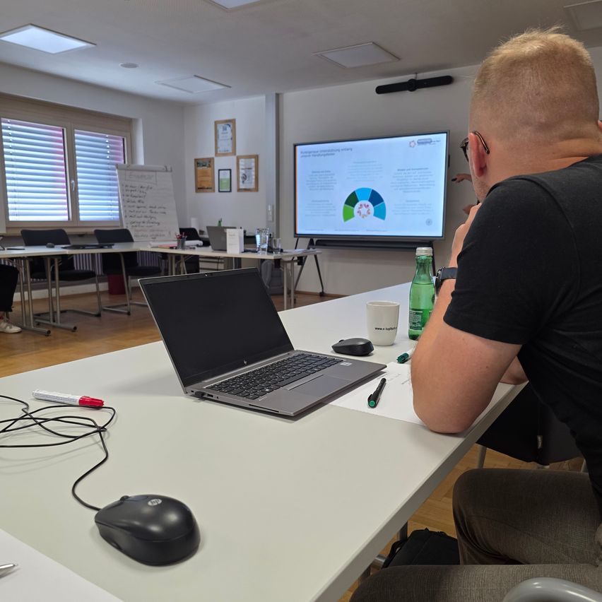 Ein Mann sitzt in einem Konferenzraum an einem Schreibtisch und konzentriert sich auf den Laptop-Bildschirm, während eine Präsentation auf einer Projektionsleinwand hinter ihm angezeigt wird.