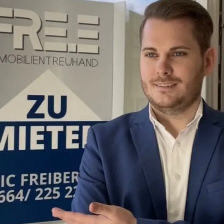 Ein Mann im Anzug steht vor einem Schild mit der Aufschrift 'Free Immobilientreuhand' und einer Telefonnummer.