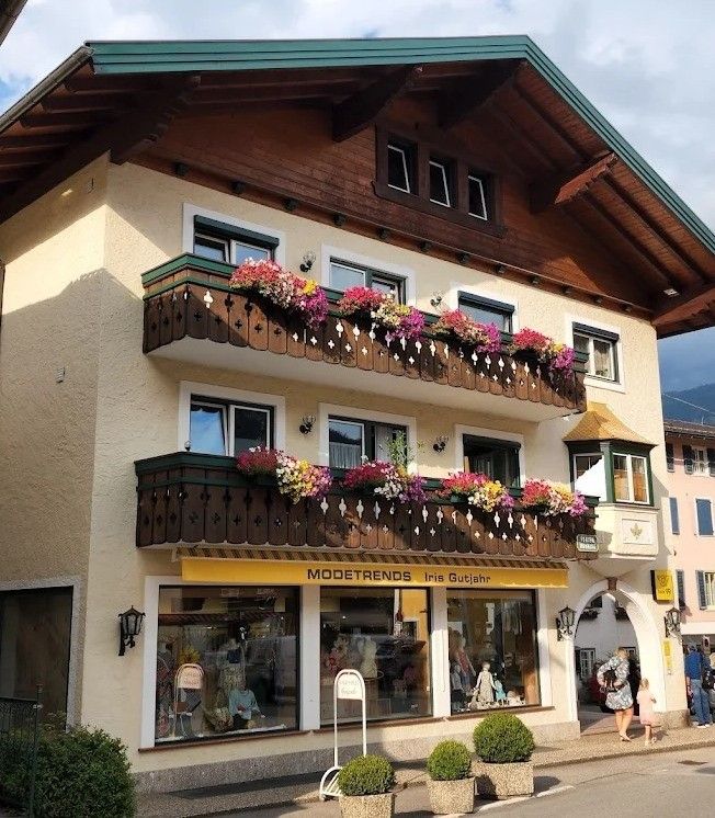 Ein Boutique namens Modetrends Iris Gutjahr befindet sich in einem mehrstöckigen Gebäude mit Balkonen voller Blumen. Eine Frau und ein Kind gehen vor dem Laden.