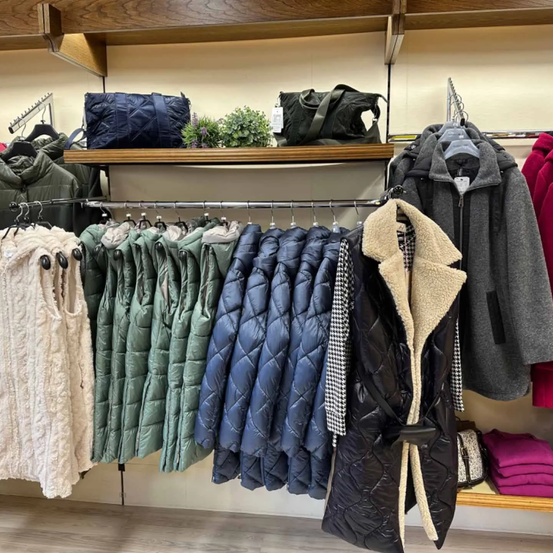 Ein Bekleidungsgeschäft mit ausgestellten Jacken und Taschen. Eine Vielzahl von Jacken hängt an Kleiderbügeln, und Taschen stehen auf einem Regal.