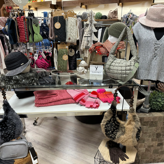 Ein Geschäft zeigt eine Vielzahl von Winteraccessoires wie Mützen, Schals, Handschuhe und Handtaschen auf Regalen und einem Glastisch. Im Hintergrund befinden sich Mannequins und gefaltete Kleidung.