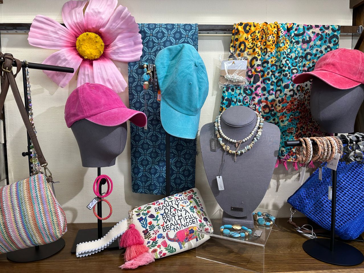 Ein Holztisch zeigt eine rosa Kappe, eine blaue Kappe, eine Handtasche, eine Tasche und Schmuck. Der Hintergrund zeigt eine große rosa Blume und einen bunten Schal.