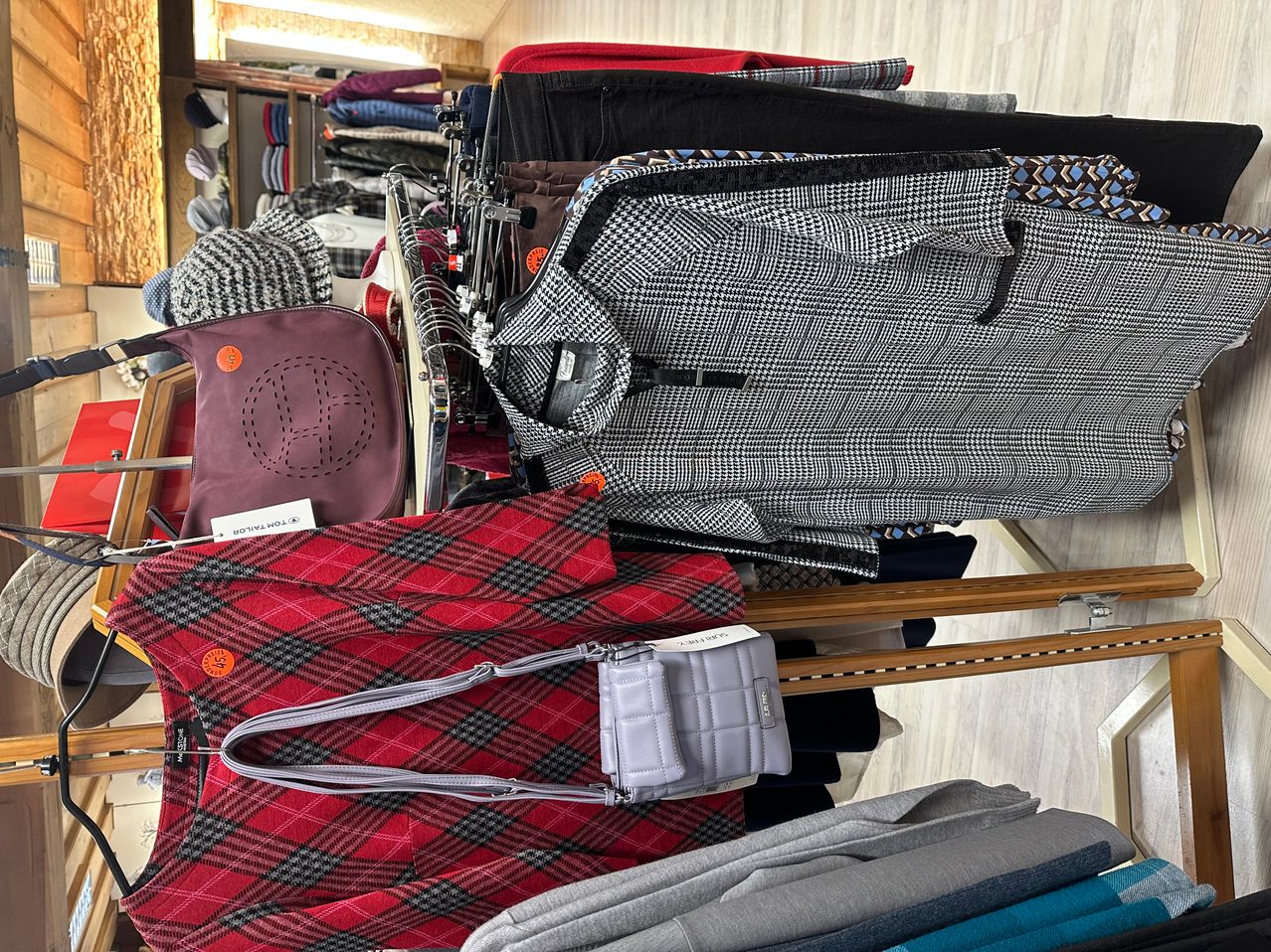 Ein Kleidungsdisplay in einem Geschäft mit verschiedenen Artikeln. Es gibt Hüte, karierte Hemden, Hosen und eine Handtasche. Die Hemden sind gefaltet und auf einem Kleiderbügel ausgestellt.