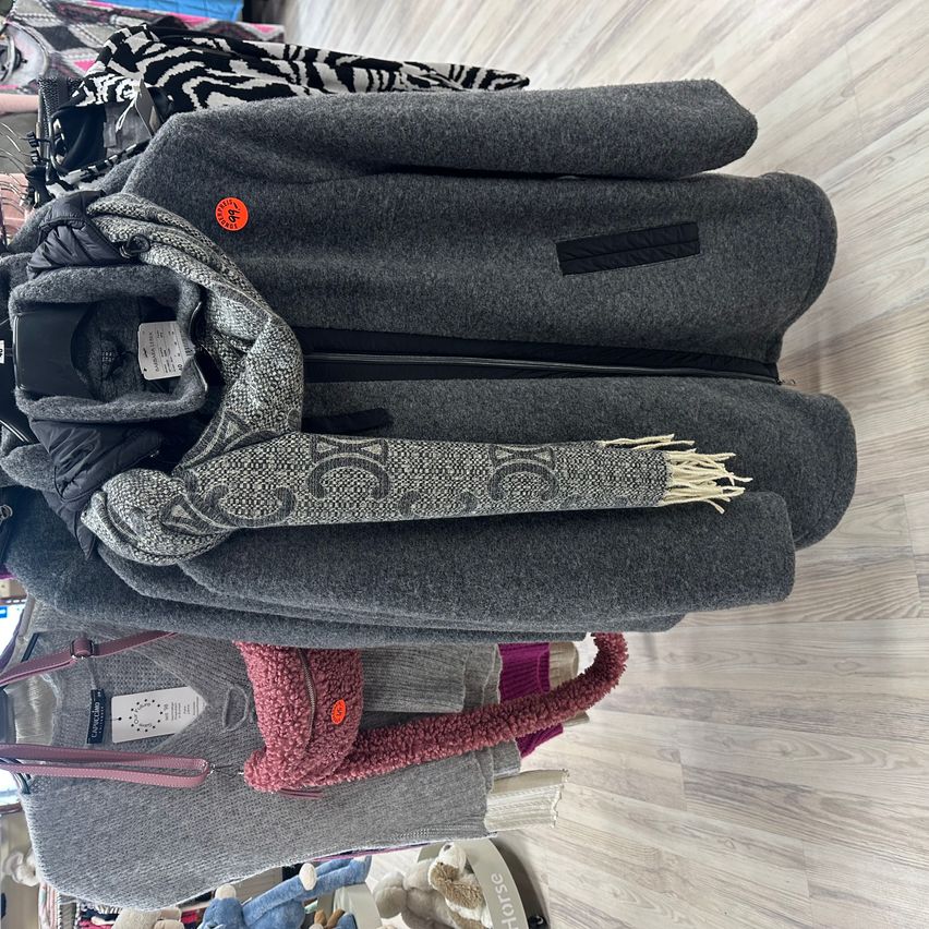 Ein Blick von oben auf einen Kleiderständer zeigt einen grauen Mantel mit einem grau-schwarz gemusterten Schal. Der Mantel hat eine Kapuze und einen Reißverschluss. Darunter befinden sich verschiedene Kleidungsstücke.