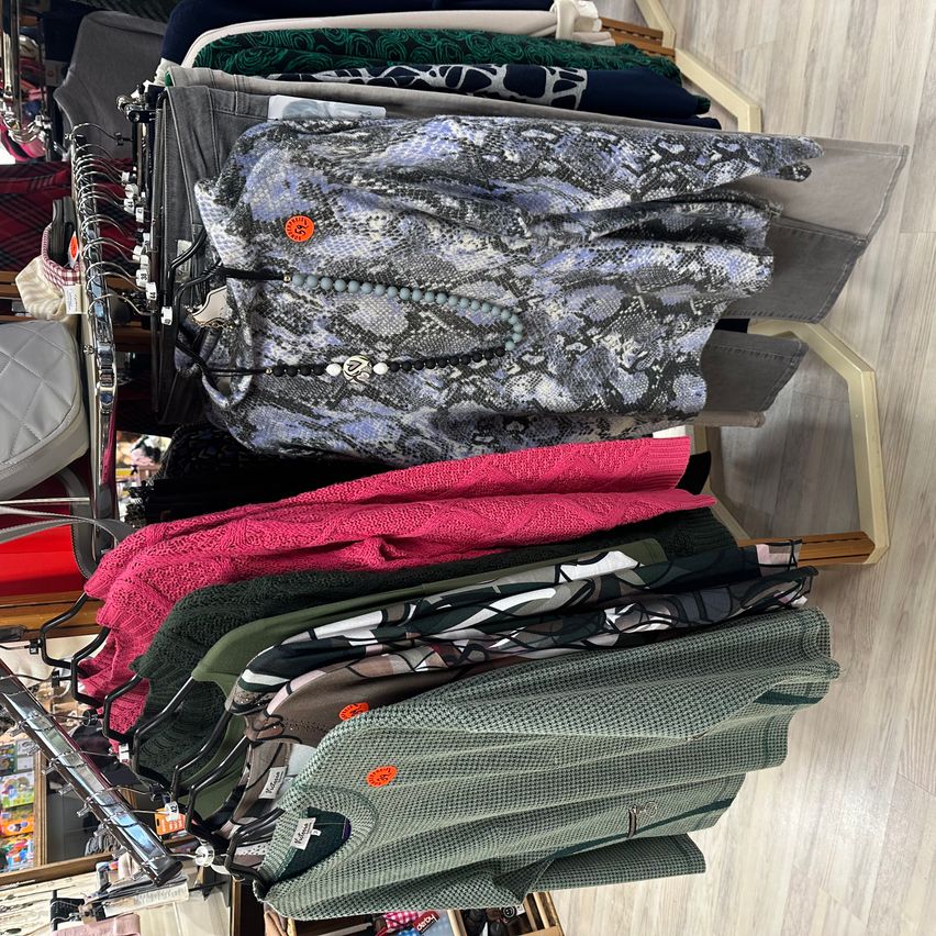 Ein Kleiderständer zeigt verschiedene Kleidungsstücke, darunter ein rosa Pullover, ein grünes Hemd und einen grauen Mantel mit Schlangenmuster. Der Ständer steht auf einem Holzboden.