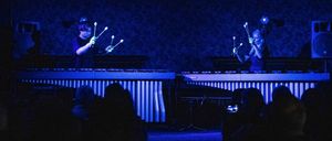 Zwei Personen spielen auf einer Bühne mit Musikinstrumenten unter blauem Licht vor einem Publikum.